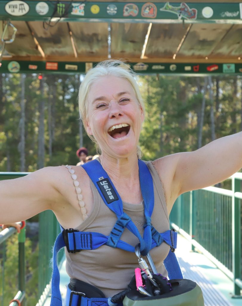A joyful woman with blonde hair wearing a harness and safety gear, smiling widely with arms raised, standing on a platform surrounded by trees.